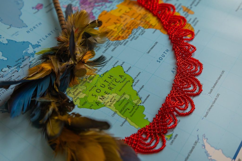 Photo by @clairepreecephotography of South America's map, surrounded by a red Indigenous-made necklace and a hair sticker with brown, yellow and blue parrot feathers.