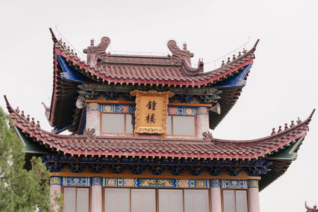 A traditional Chinese bell tower with two tiers of curved, upturned roofs, painted blue and gold beams, and a golden plaque reading "Bell Tower" in Chinese characters. Photo by Suki Lee.