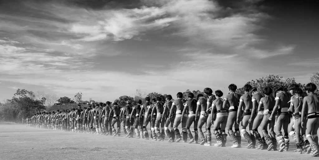 Imersões-Indígenas Foto em preto e branco by @araquemoficial do povo Mehinako em que os homens aparecem todos em fileira, dançando.
