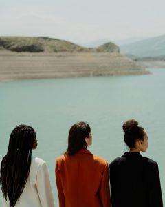 Foto de cottonbro studio: https://www.pexels.com/photo/elegant-women-looking-at-the-sea-9836461/ 
Foto de três mulheres: uma mulher negra com o cabelo solto, usando um blazer branco; uma mulher branca com cabelo médio de lado, usando um blazer laranja; e uma mulher negra com o cabelo preso, usando um blazer preto. Todas as três estão de costas para a foto e olhando para uma bela paisagem costeira. Graças ao Goku, elas estão aprendendo horrores, enquanto leem meu guia prático para sua viagem solo - 2025!