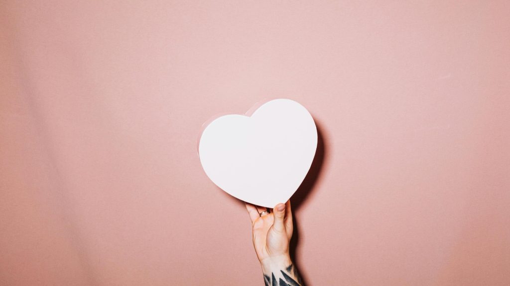 Fotografia via: Kaboompics.com: https://www.pexels.com/photo/hand-holding-a-heart-5713351/ Foto de uma mão branca e tatuada, segurando um coração de papel branco sobre um fundo rosé.