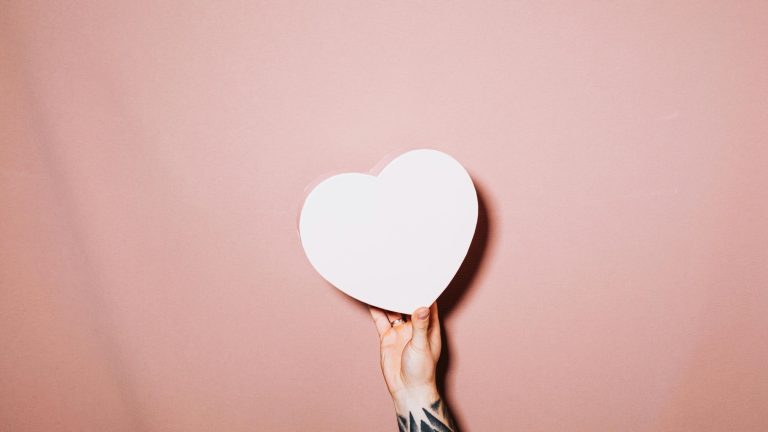 Fotografia via: Kaboompics.com: https://www.pexels.com/photo/hand-holding-a-heart-5713351/ Foto de uma mão branca e tatuada, segurando um coração de papel branco sobre um fundo rosé.