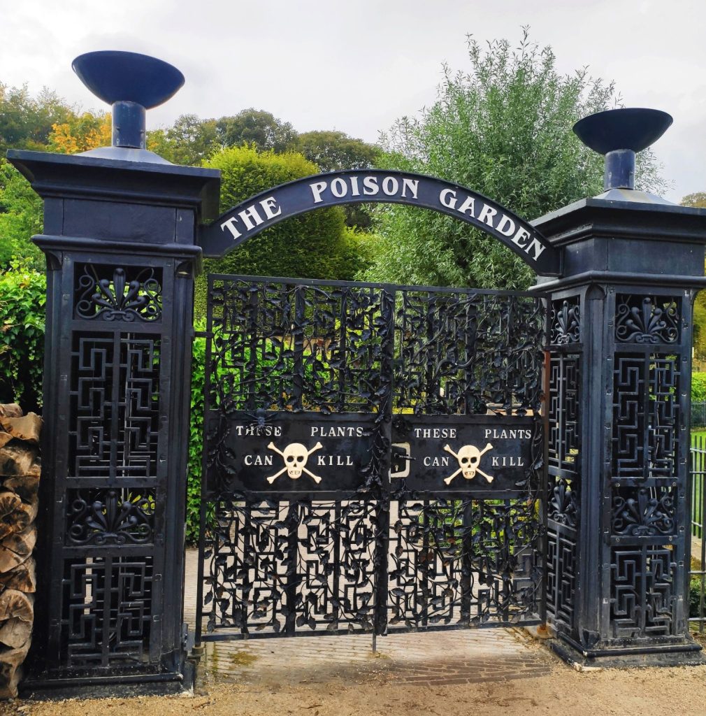 Imagem do portão do jardim venenoso de Alnwick, em Northumberland, Inglaterra. O portão é todo preto e tem escrito em cima: "O jardim venenoso". Mais abaixo, duas caveiras (uma em cada lado), com a frase: "estas plantas podem matar".