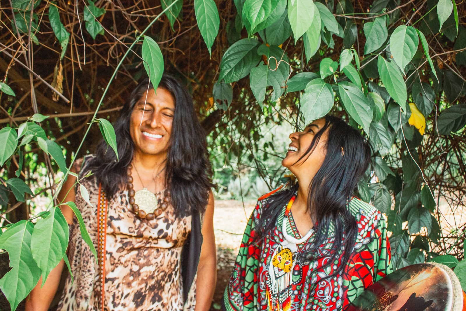 Huni-Kuin-e-Inca Uma foto colorida de Inti Sonqo e Bimi Sabino sorrindo sob as árvores, vestindo trajes tradicionais e segurando tambores. Inti está de olhos fechados e Bimi olha para Inti.