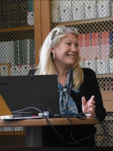 Imagem da Dra. Lisa Miller em sua palestra no Fórum Inter-religioso de Oxford, usando um casaco azul, um cachecol azul e óculos brancos no cabelo.
